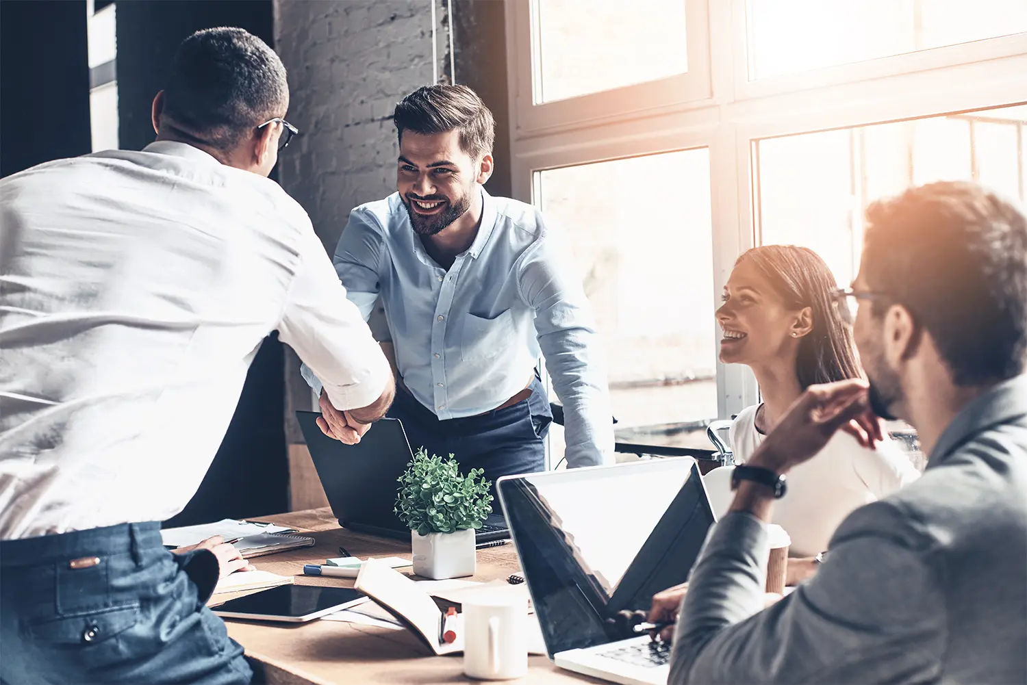 Four employees sitting at a table, two shaking hands while the other two smile at them.